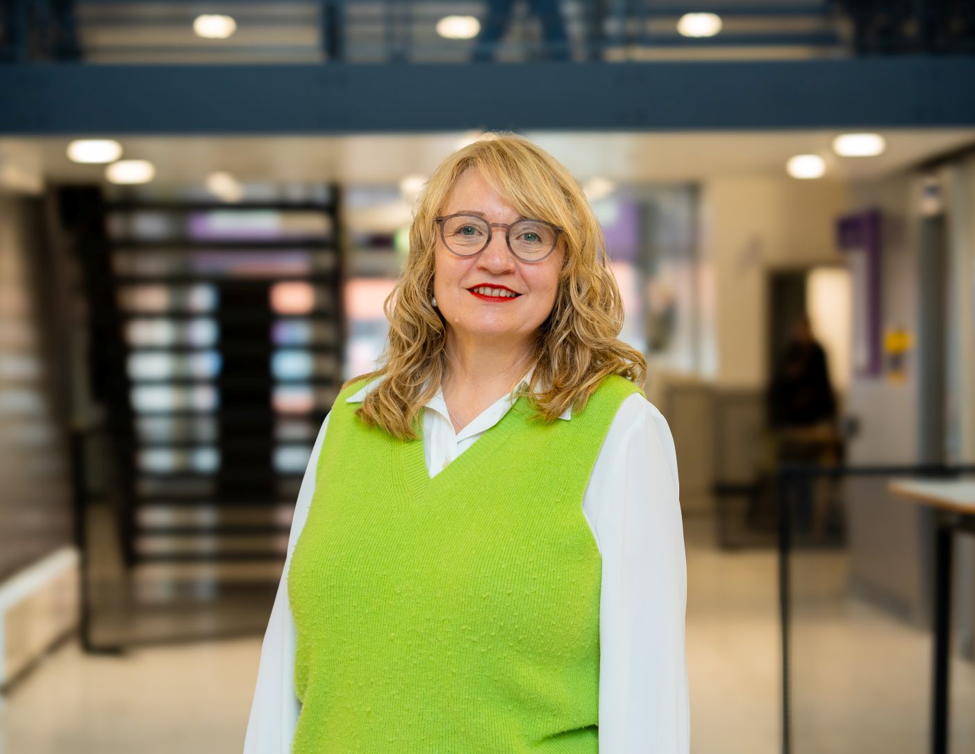 Image of Liz Birchinall. A female academic with blonde hair, wearing red lipstick and a green vest. 